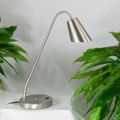 Amsterdam bordlampe tændt på et bord mellem grønne planter, sølvfarvet skærm og rund fod