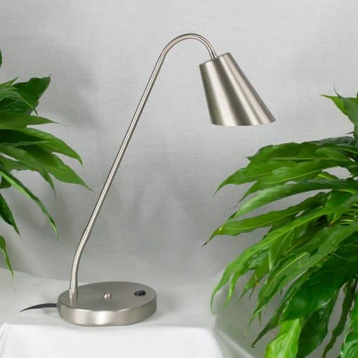 Amsterdam bordlampe tændt på et bord mellem grønne planter, sølvfarvet skærm og rund fod